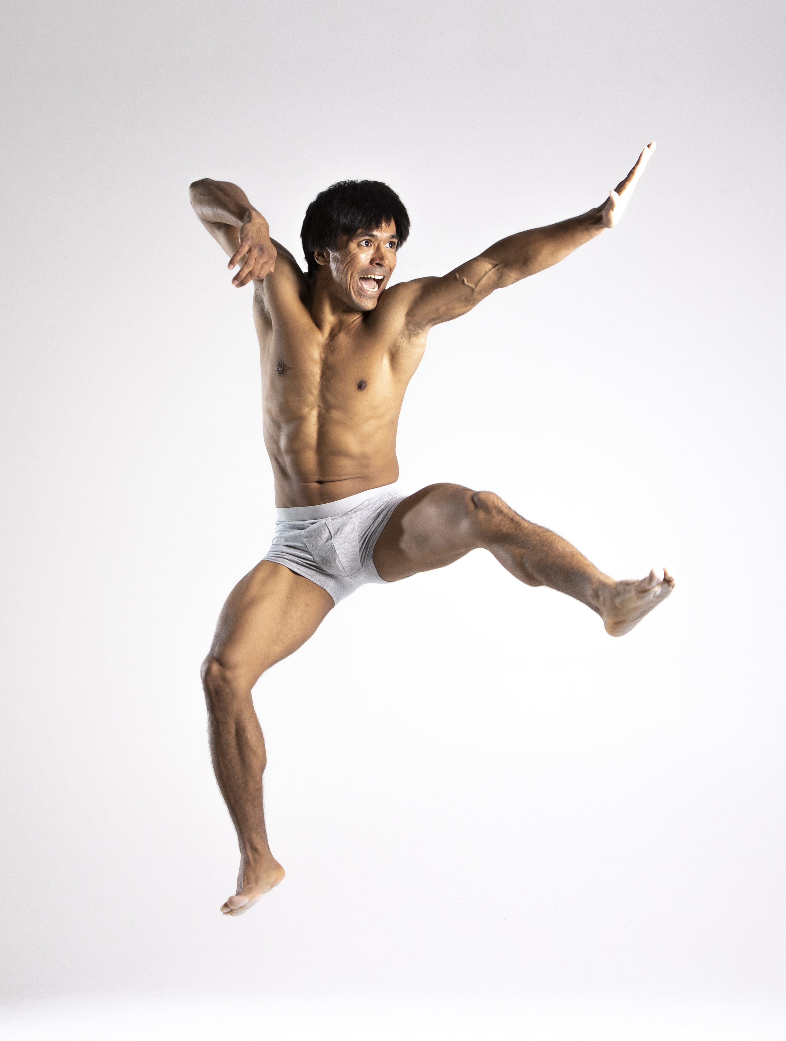 a muscular, shirtless asian man is captured midair in a dynamic jumping pose. he is wearing light grey underwear and is barefoot. his body is tense with energy as he expresses excitement or action, suggesting fitness, strength, or movement such as dance or martial arts. the background is plain white, isolating the man as the focal point and emphasizing his physique and movement.
