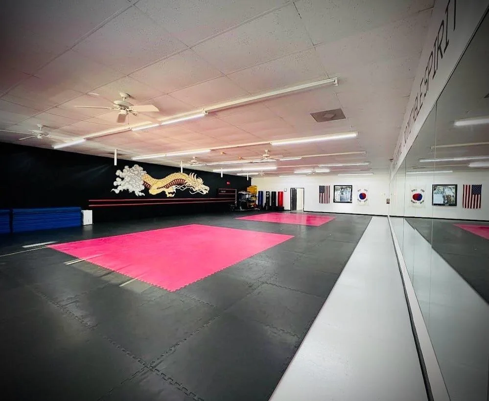 an indoor martial arts gym with a large pink matted area in the middle of the room. the walls are white and the ceiling is high with several fluorescent lights. the room features large mirrors on one wall and a black wall with a gold dragon mural on the opposite wall.