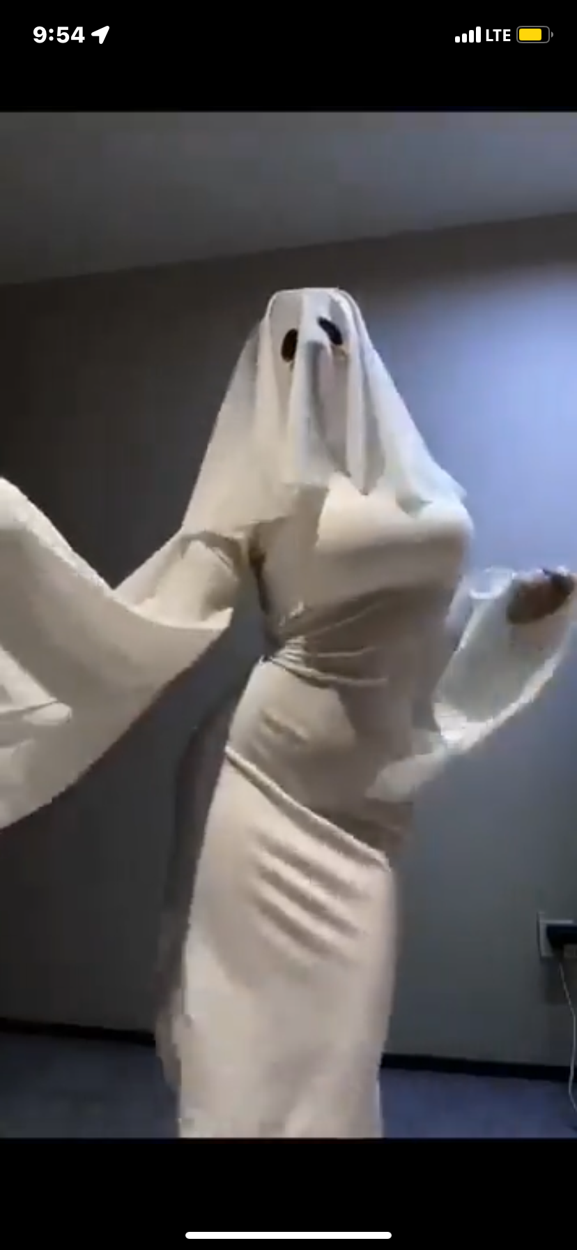 a woman is dressed up in a ghost costume. she is wearing a white dress and has a white sheet over her head.  she is standing in a room and it is dark.