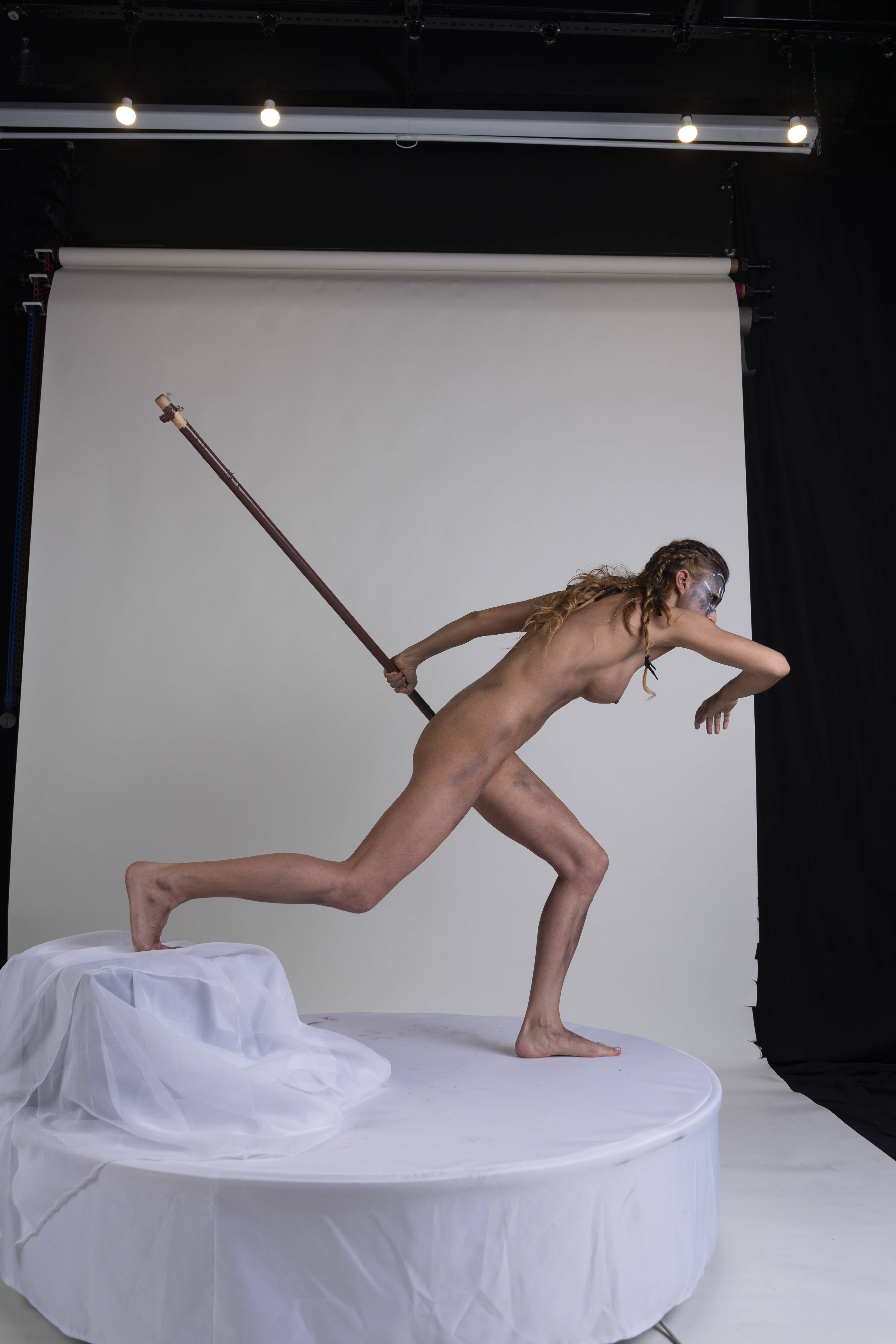 a nude woman stands on a white platform, with one leg raised as if running. she holds a staff behind her and wears face paint. the background is a studio setting with a white and black backdrop.
