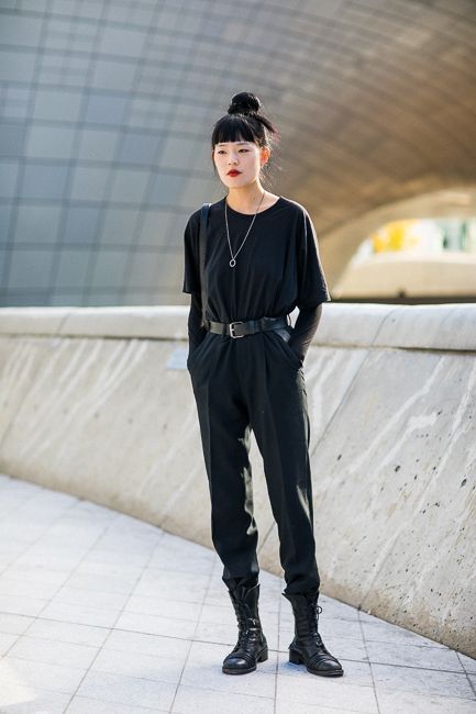the image shows a young asian woman showcasing street style fashion. she is wearing a black jumpsuit with a slightly loose fit and long sleeves, cinched at the waist with a black belt.  she's paired the jumpsuit with black combat boots that reach her midcalf.  her hair is styled in a high bun, and she wears a simple silver necklace. the backdrop is a modern, concrete architectural structure, suggesting an urban setting.  the overall style is minimalist and slightly edgy, emphasizing dark colors and clean lines.  the woman appears confident and stylish in her outfit.