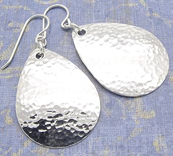 a pair of silver teardropshaped earrings with a hammered texture are displayed on a blue textured surface. the earrings have a shiny finish and are hanging from a silver hook.