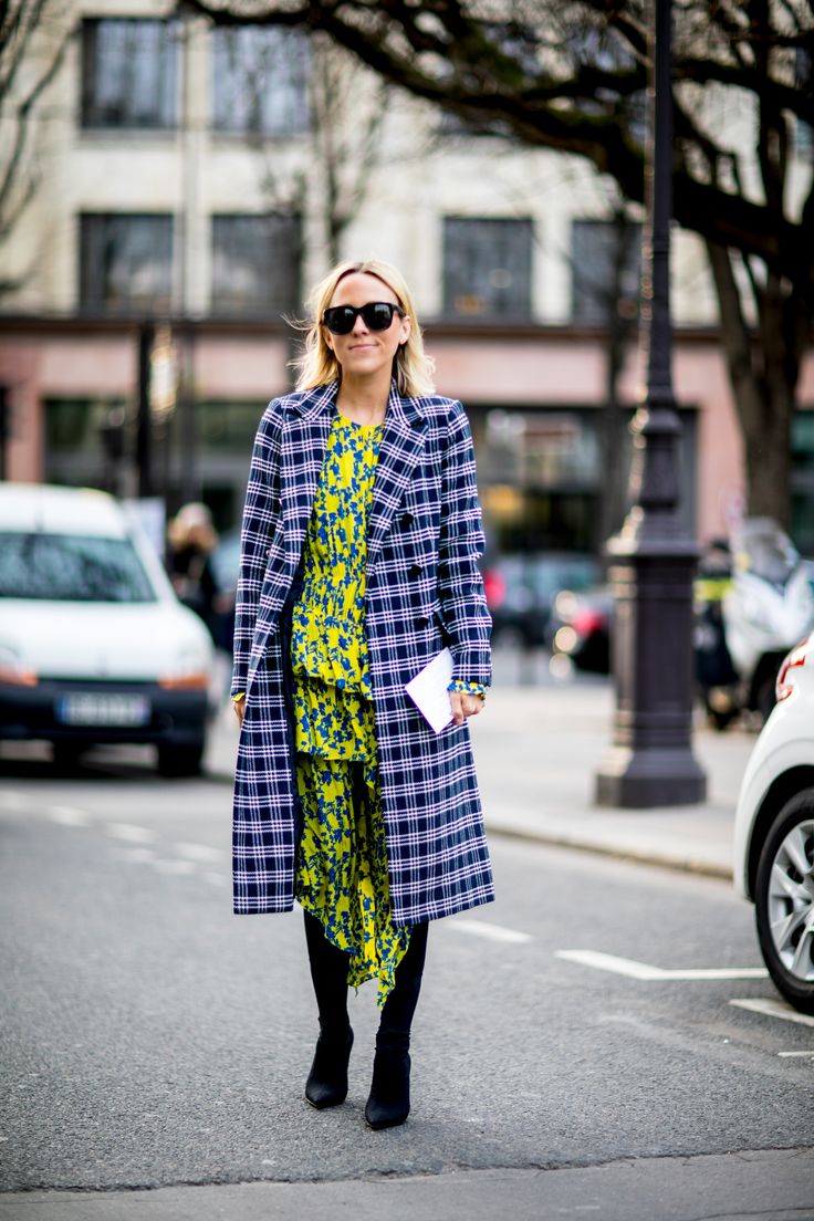 a stylish woman in a plaid coat and yellow floral dress is walking down a city street. she is wearing black ankle boots and sunglasses. the background is blurred but shows a city street with cars and trees. the overall style is chic and elegant.