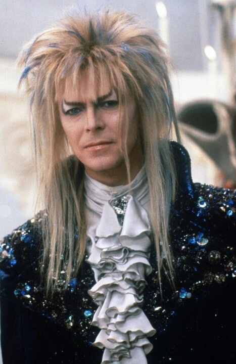 a portrait of david bowie as jareth the goblin king from the 1986 movie labyrinth. he is wearing a blue and black velvet jacket with sequins and jewels. he has a white ruffle collar and white makeup.  he has very long blonde hair.  he is looking into the camera.  his expression is serious and intense.