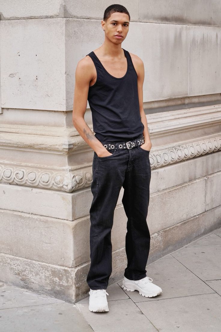 a young male model stands against a stone wall, showcasing a casual yet stylish outfit. he's wearing a black tank top, darkwash black jeans, a studded belt, and white chunky sneakers. his pose is relaxed, and his expression is neutral and confident. the background features a textured stone wall, typical of an urban environment. the overall aesthetic is contemporary and urbanchic.