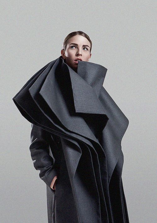 a fashion model wearing a grey coat with a very large, geometric collar. the model is posing in a studio setting, against a grey backdrop. the model is looking up towards the left. the overall look is sleek and modern, with a strong emphasis on the unique design of the coat.
