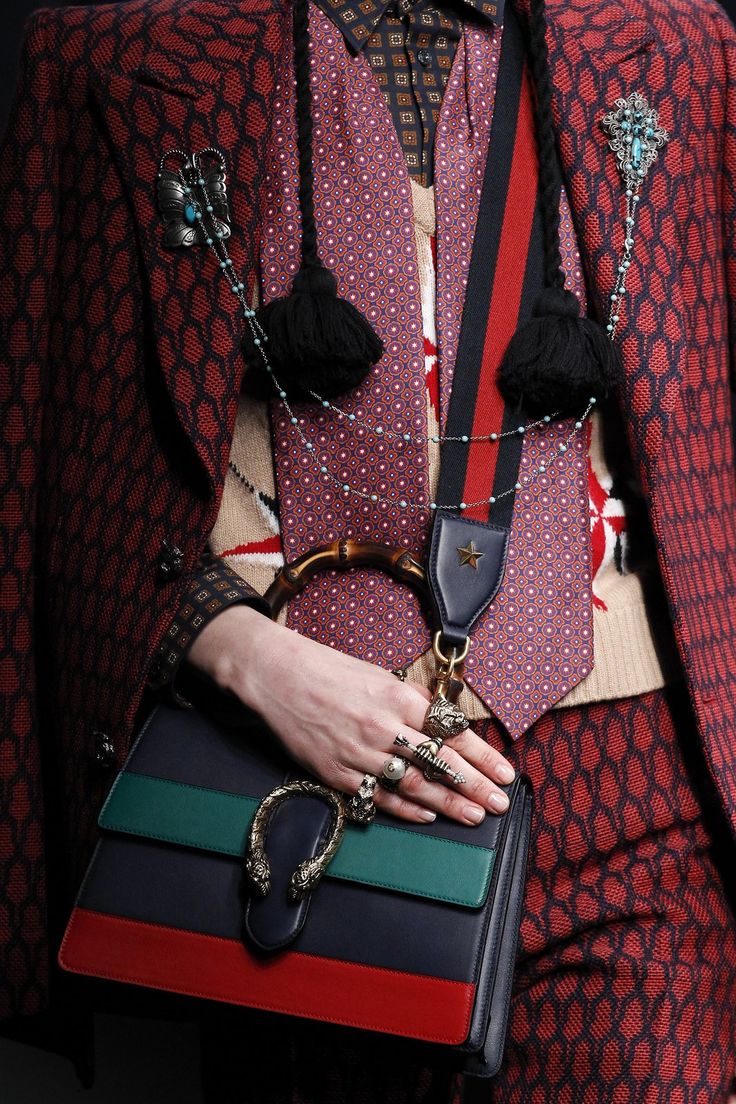 closeup shot of a model showcasing a gucci outfit. the focus is on the accessories: a structured handbag with a distinctive gucci horsebit detail, layered necklaces with brooches and tassels, and multiple rings. the model is wearing a red patterned blazer, a patterned tie, and a beige sweater. the image highlights the intricate details and luxurious textures of the garments and accessories.