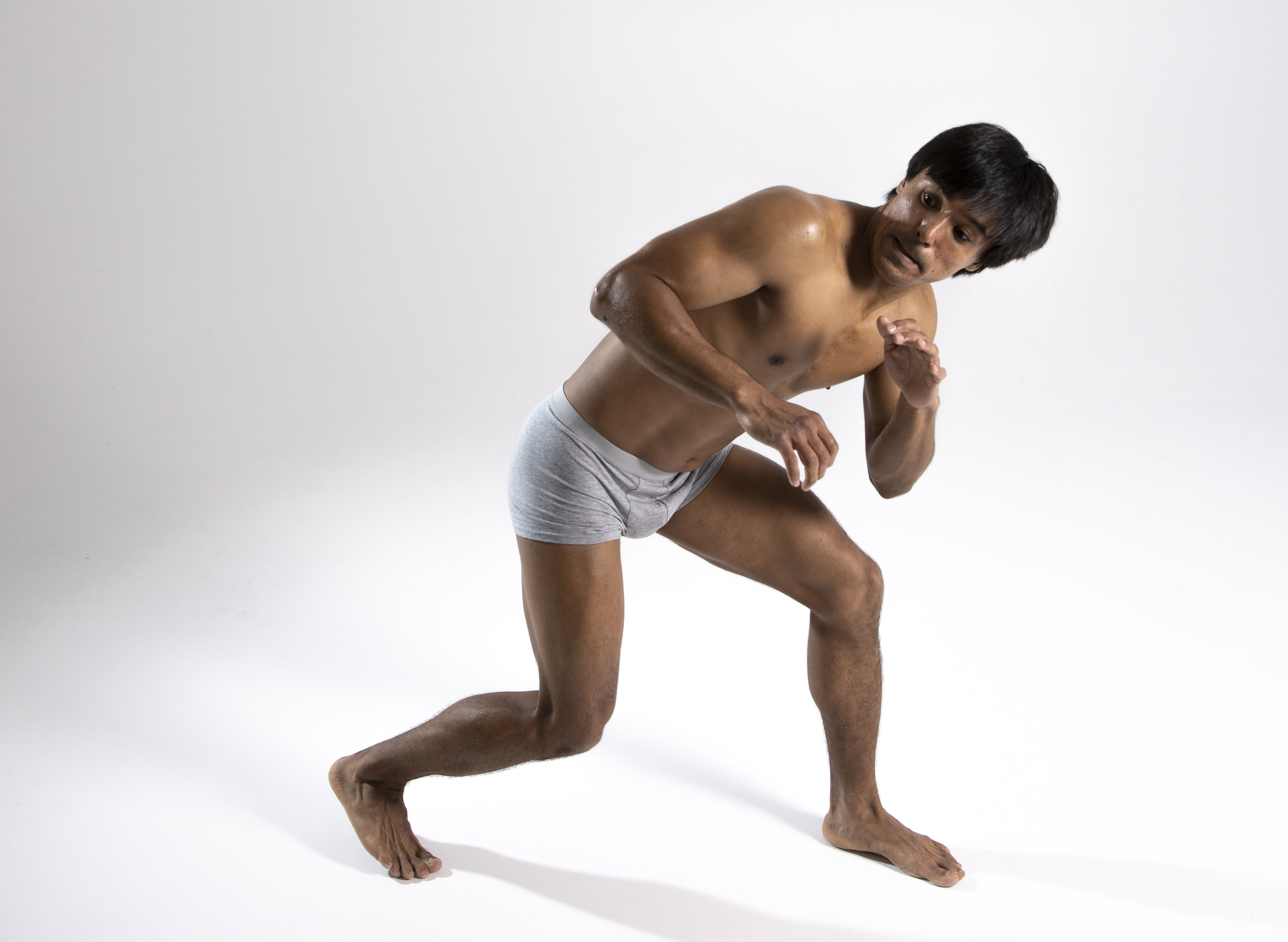 a full shot of a muscular, darkskinned man in gray underwear, posing barefoot on a light background indoors. he is crouching defensively with a scared expression, as if dodging or avoiding something. the man is looking at the camera, displaying surprise and a startled gesture. the image is a studio shot with bright lighting and a white background, emphasizing the man's athletic build and physique.