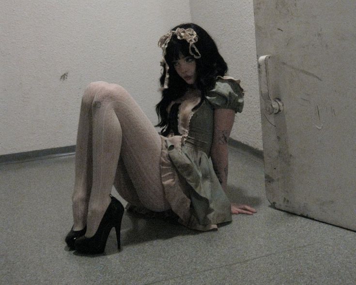 a woman is sitting on the floor wearing a dress, stockings, and high heels. she has tattoos and a flower crown on her head. the background consists of a wall and a door.