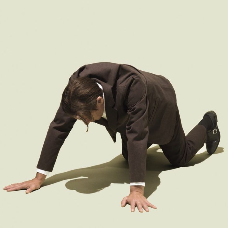 a man in a suit is crawling on the floor. he is looking down and his face is serious. the background is a light green color. he looks very stressed, almost helpless, as if he has failed.