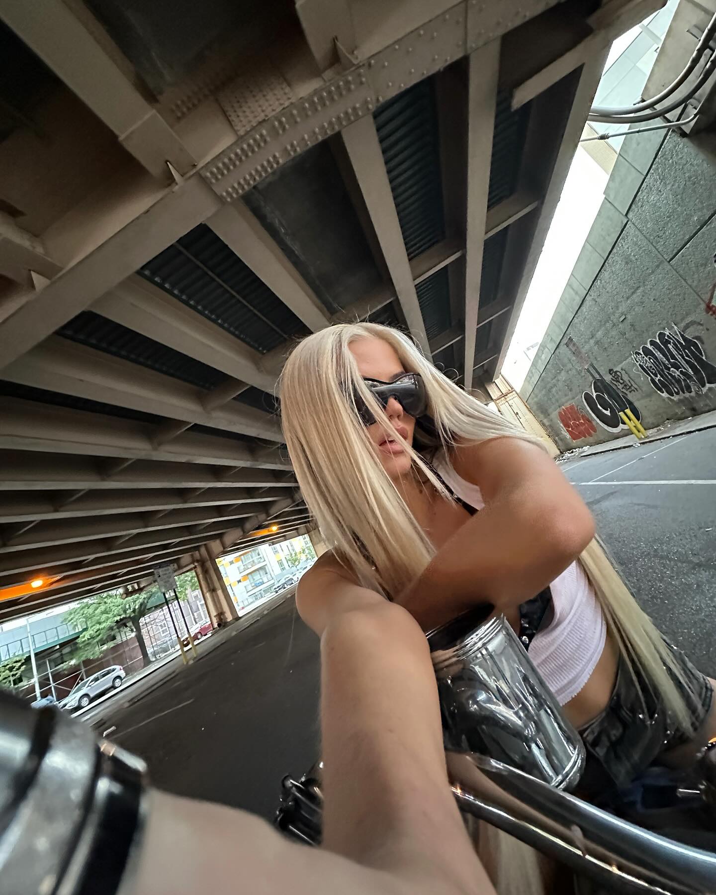 a woman with long blonde hair and sunglasses is sitting on a motorcycle under a bridge in an urban setting. there is graffiti on the wall behind her. buildings are visible in the background.