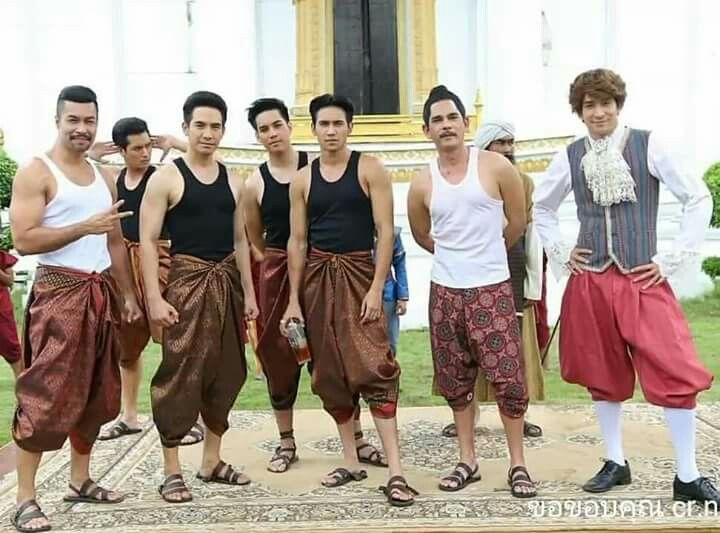 a group of seven men are standing together in a garden setting. they are all wearing traditional thai clothing. the men are smiling and appear to be happy and relaxed. they are all dressed in bright, colorful clothing that would be considered formal attire. the setting is outdoors and the background appears to be a palace or temple. the group is a mixture of young and older men. this group is likely from thailand and is wearing traditional clothing for a special event or occasion.