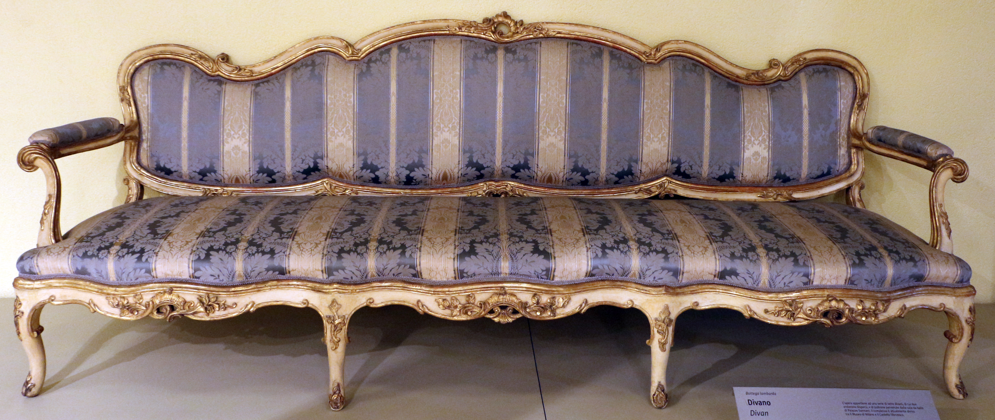 a rococostyle divan or couch with ornate, gilded frame and striped, upholstered cushions in blue and gold tones, sitting in a room.