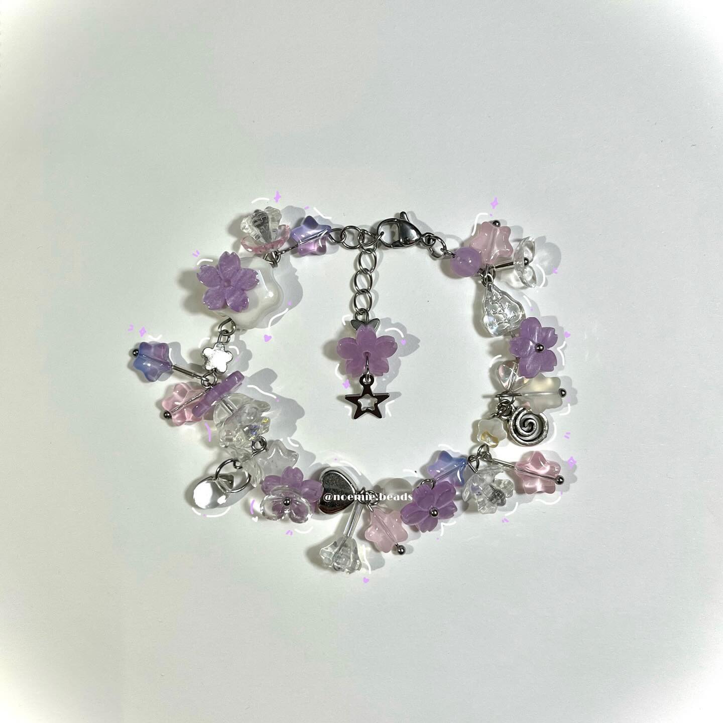 a beaded bracelet is centered on a white background. the bracelet is made of a silver chain with various clear, light pink, and light purple beads and flower beads. a star charm hangs from one of the flower beads. the text "@nceonie.beads" is also on the bracelet.