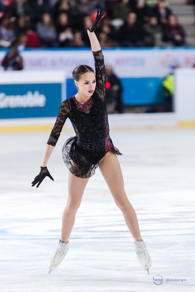 a female figure skater in a black dress is skating across the ice. she is performing a move where she has one leg extended high in the air. her other leg is on a skate with the skate blade digging into the ice and causing a blurry effect. the athlete is focused and in motion, as she performs a dynamic and graceful move on the ice.