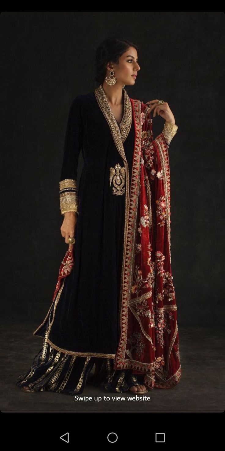 the image showcases a woman elegantly dressed in a dark navy blue velvet sharara suit paired with a richly embroidered maroon dupatta. the sharara is adorned with gold embroidery along the hem and neckline. the dupatta features intricate floral patterns and gold detailing along its borders.  she is wearing gold earrings and is posed against a dark backdrop, creating a dramatic and sophisticated aesthetic. the image has a promotional feel, likely for a fashion brand, as indicated by the text "swipe up to view website."