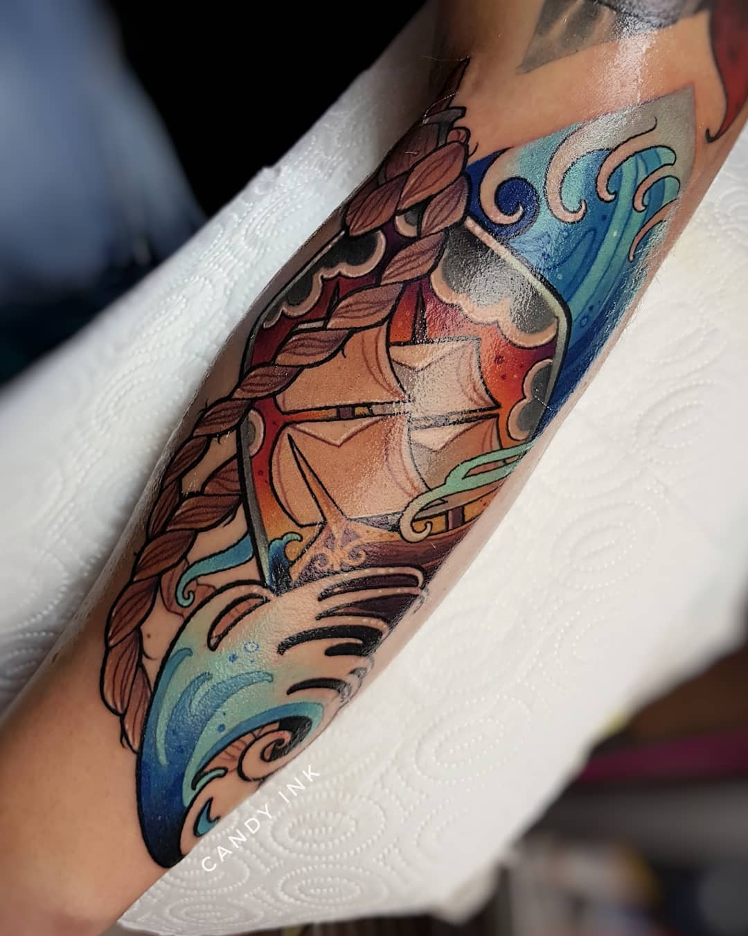 a closeup photograph of a colorful neotraditional tattoo of a ship on someone's arm. the tattoo is of a ship sailing on the ocean with clouds in the sky. the tattoo is surrounded by a rope border. the tattoo is done in a traditional style with bold lines and bright colors. the person's arm is resting on a white surface.