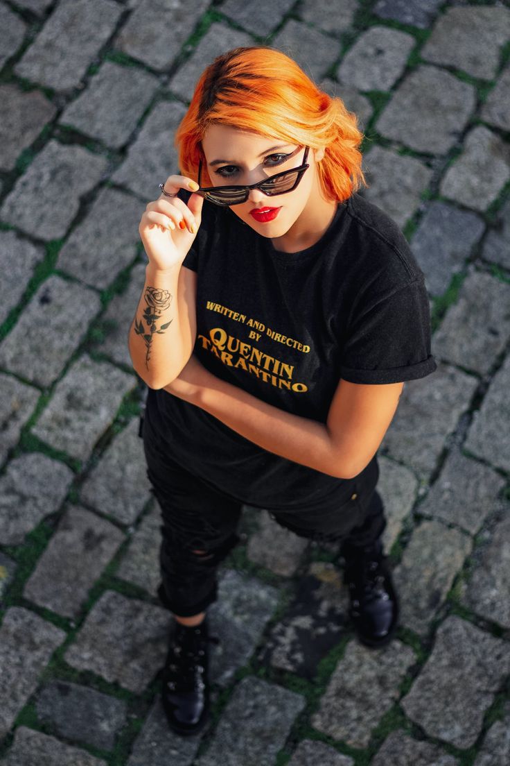 a young woman with bright orange hair and black sunglasses is standing on a cobblestone street. she is wearing a black tshirt with the words "written and directed by quentin tarantino" printed on it, black ripped jeans, and black boots. she has a rose tattoo on her left arm.
