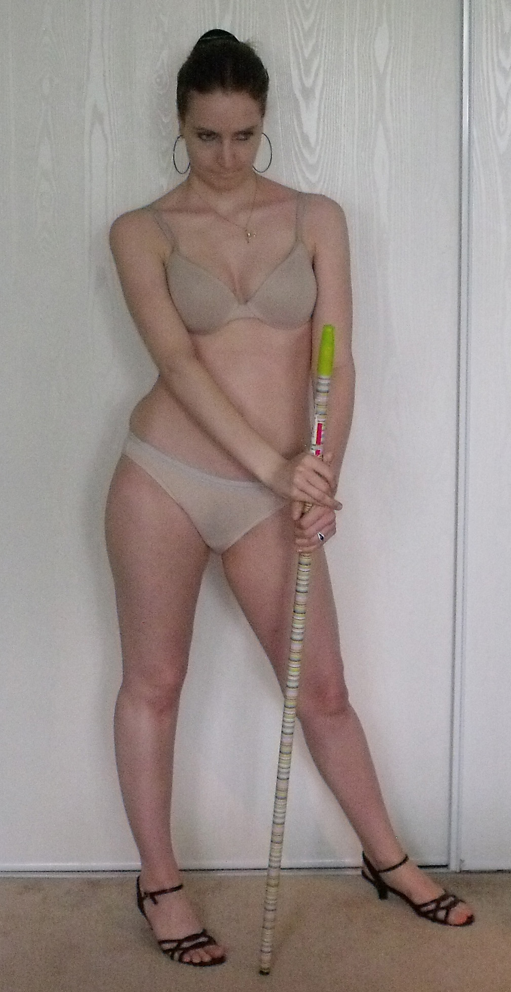 a fullbody shot of a woman standing in lingerie, holding a long, thin stick. she's wearing a beige bra and panties, and black heels.  her expression is neutral, and she's standing against a lightcolored wooden wall. the image appears to be a studio shot.