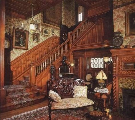 the image shows a grand victorian era wooden staircase in an opulent home interior.  the stairs are carpeted in a rich red, and the walls are covered in patterned wallpaper. a variety of antique furniture, including a chaise lounge and side table, are arranged near the staircase and fireplace. a warm, inviting light fills the space, highlighting the intricate carvings and rich textures.
