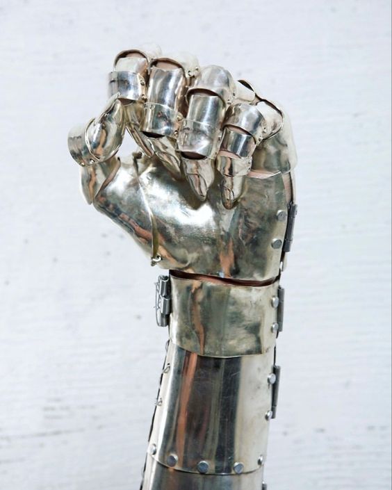 a closeup image of a sculpted metal arm and hand. the hand is formed into a fist and is highly detailed, with individual plates of metal mimicking the structure of armor. the metal has a polished, silver finish, and the piece appears to be photographed against a neutral, textured background.