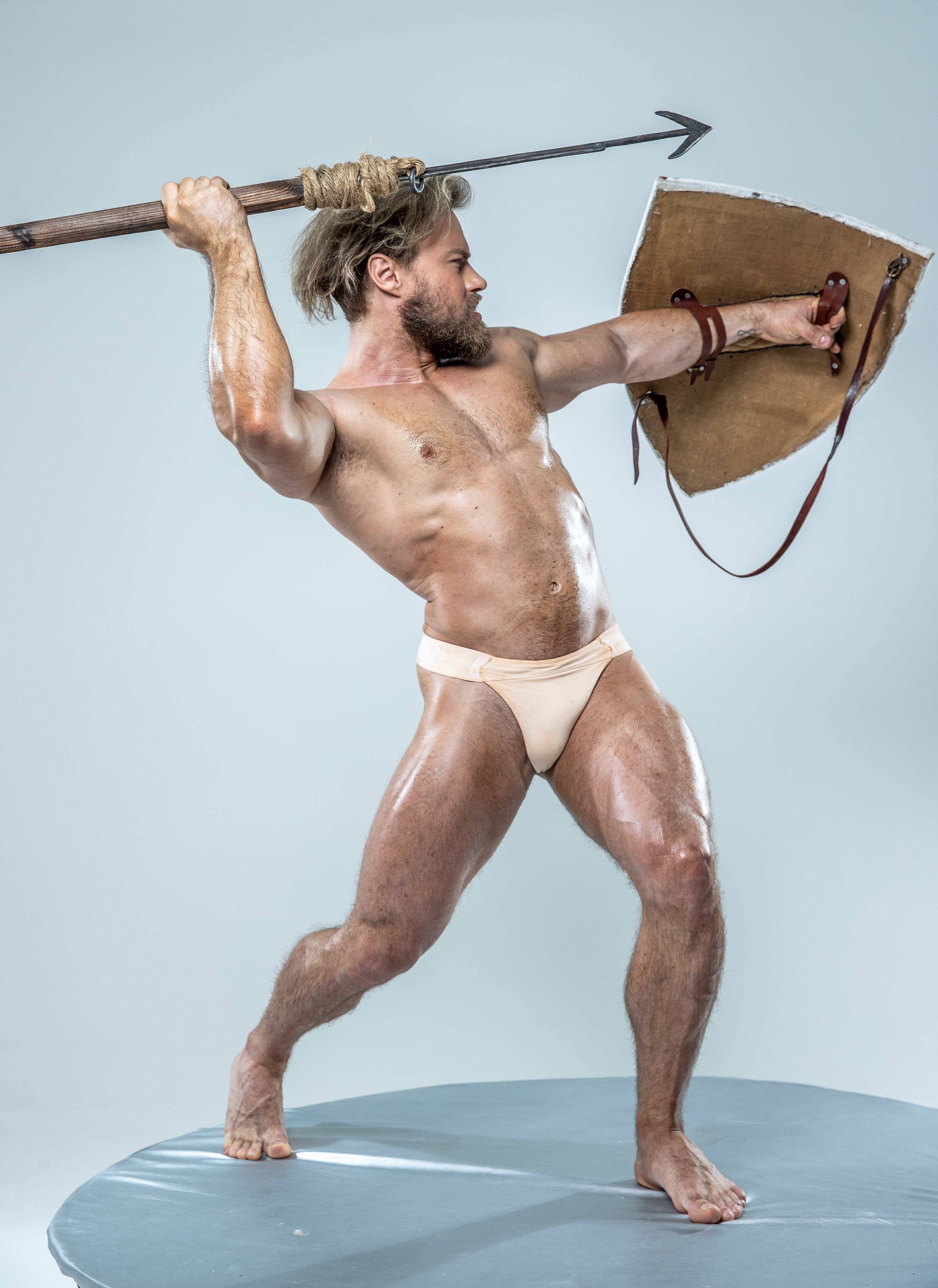 a muscular, shirtless man with long, braided blond hair stands on a circular platform against a light gray background. he is posed dynamically, wielding a spear with a barbed metal point in his right hand and a round leather shield in his left. he appears to be in the act of attacking, his body tensed with power. his attire is minimal, consisting only of a beige loincloth. the studio lighting highlights his muscular physique and creates a sense of action in the image.