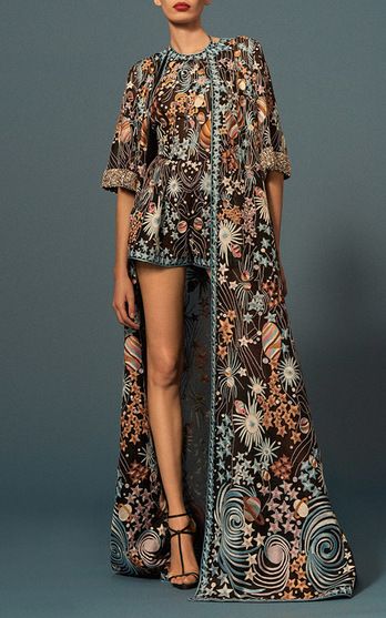 a model is showcasing a stunning, highlow embroidered dress. the dress features a short jumpsuitstyle top with a long, sheer overlay. the embroidery is intricate and detailed, with floral, star, and galaxylike patterns in a muted color palette.  the model wears highheeled sandals and poses against a dark blue backdrop. the overall style is elegant and sophisticated.