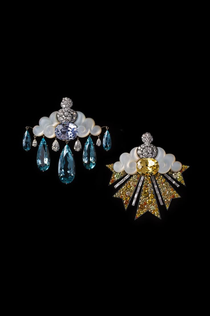 two pieces of luxury jewelry are showcased against a black background. both pieces feature a cloudlike base crafted from white material, possibly motherofpearl, adorned with diamonds and gemstones. the left piece has a large blue gemstone in the center with teardropshaped blue pendants dangling below, while the right piece has a central yellow gemstone and radiating rows of yellow and clear gems.