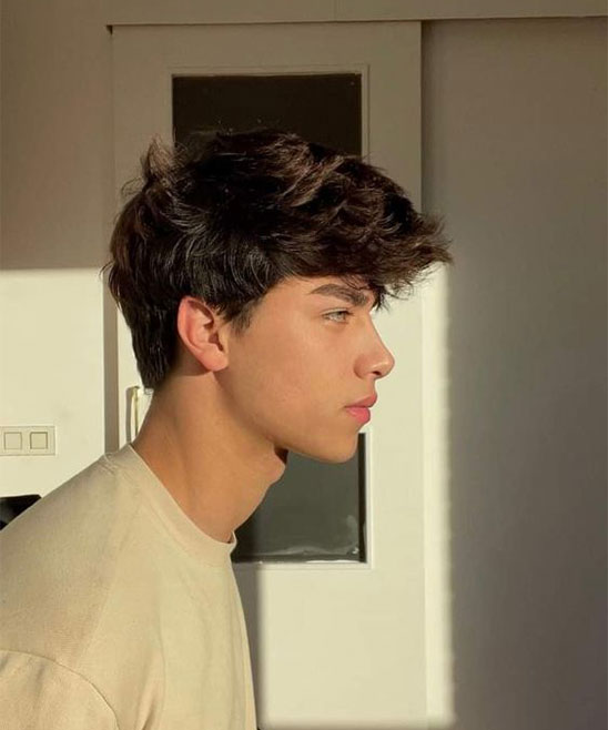 a young man with short brown hair is posing in a light cream shirt,  looking to the side. his expression is neutral.  he is standing indoors with a light background and a shadow from a window. the man is slightly turned to the right.  his hair is styled neatly and slightly curled.  his eyes are brown, and his skin is pale.
