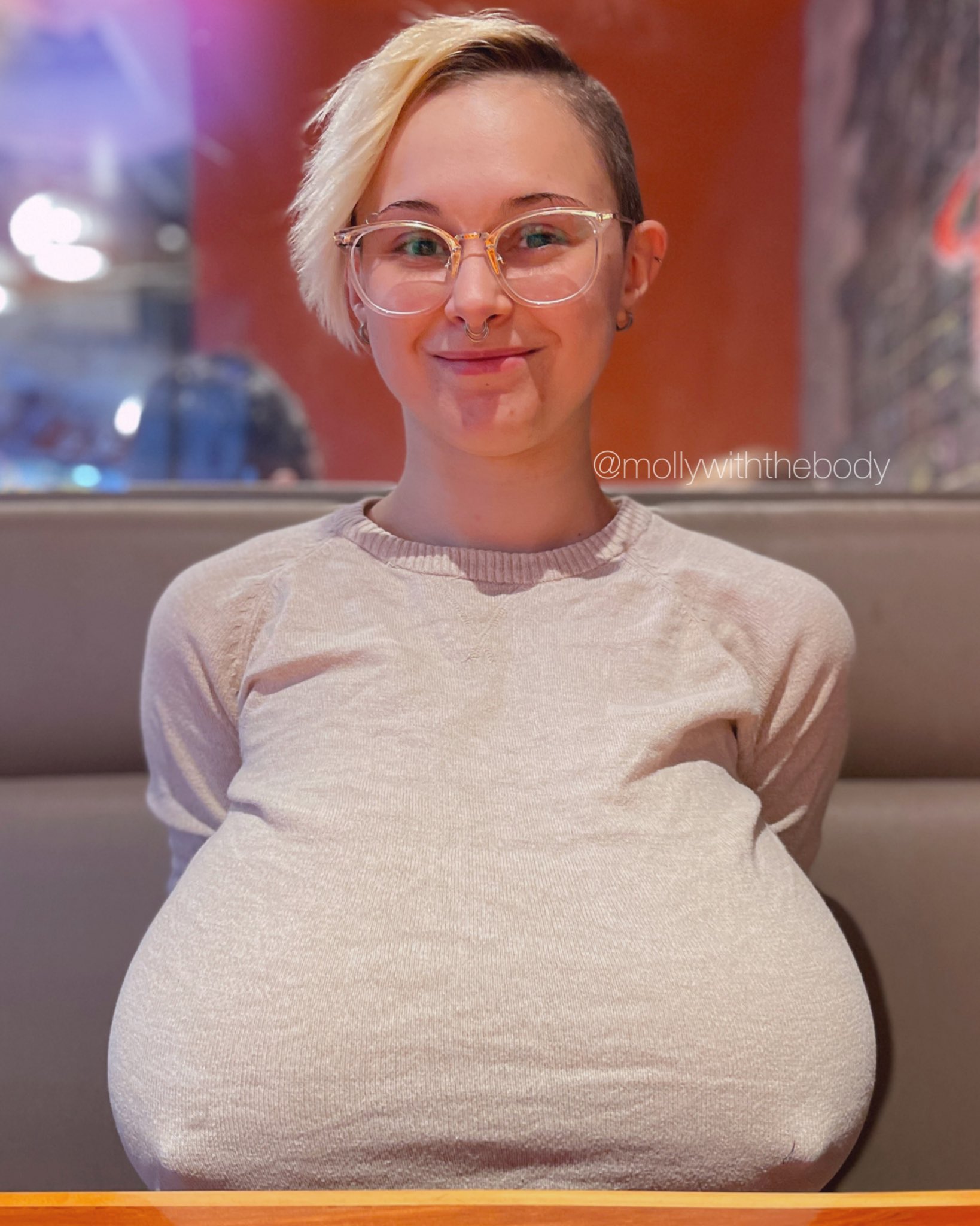 a closeup portrait of a smiling young woman with short blonde hair and clear glasses.  she's sitting in a restaurant booth wearing a light beige sweater that accentuates her large breasts. the overall impression is one of confidence and body positivity.  an instagram handle, @mollywiththebody, is visible in the image.