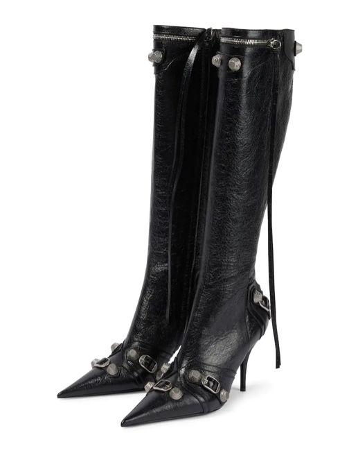 the image shows a pair of balenciaga women's kneehigh boots.  they are black leather with a pointed toe and stiletto heel. the boots are adorned with silvertoned studs and buckles along the shafts and straps. the boots are positioned angled, showing the profile of both boots, highlighting the detailing and the shape of the heels.