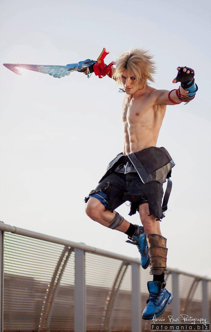 a man dressed as the character cloud strife from the final fantasy series is jumping in the air, holding a sword. he is wearing a costume that includes a blue shirt, black shorts, and blue boots. the background is an urban scene with a metal fence in the foreground. the image is likely a cosplay photograph.