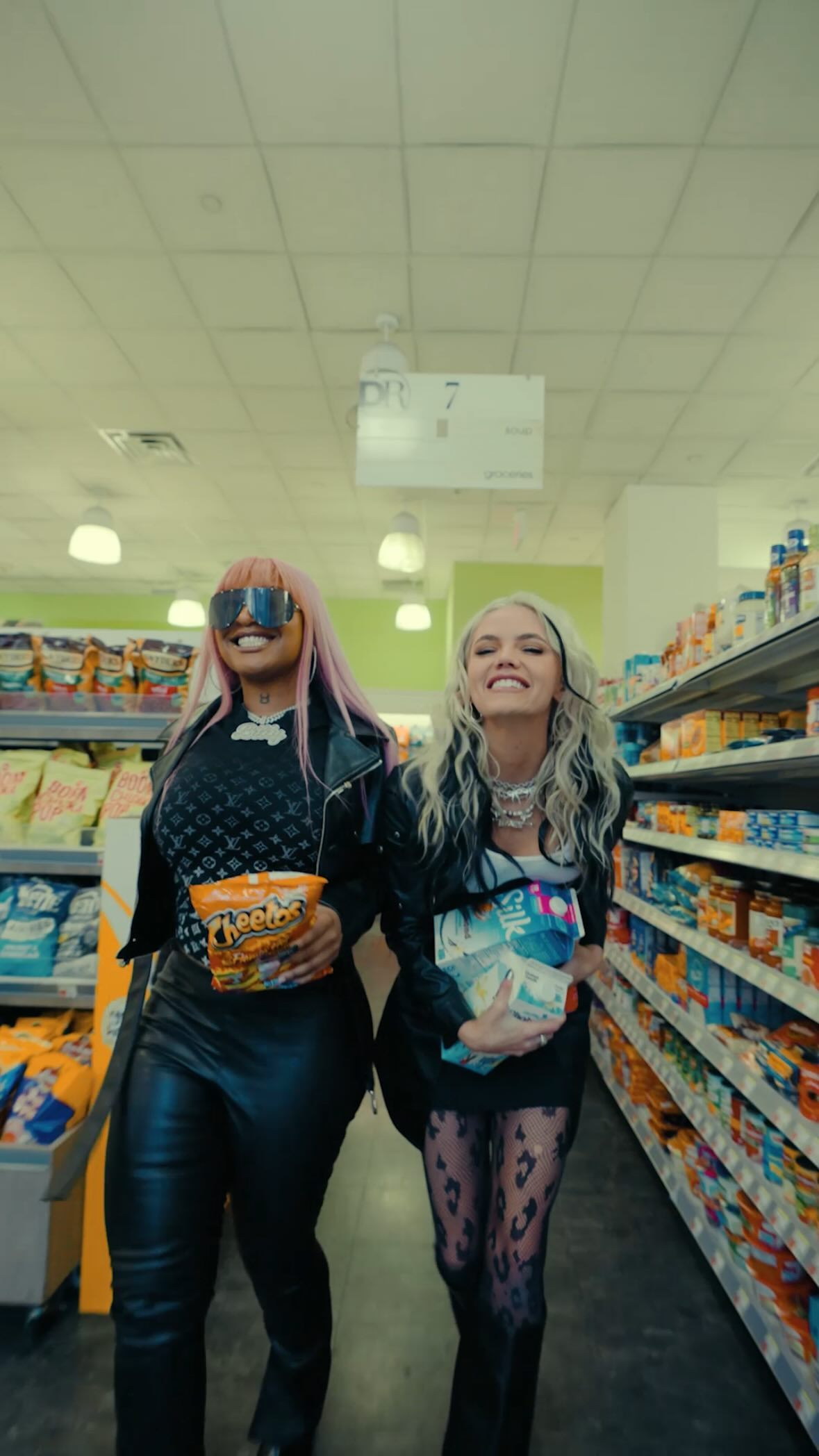 two women are walking through a grocery store aisle. the woman on the left has pink hair, sunglasses, and a black leather jacket, and she is holding a bag of cheetos. the woman on the right has blonde hair and a black leather jacket, and she is holding a package of silk plantbased milk. they are both smiling and appear to be enjoying their shopping trip. the background shows aisles filled with various grocery products.