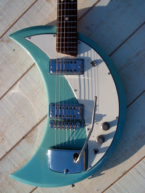 a closeup view of a blue and white crescentshaped electric guitar. the guitar has a unique design, with a curved body that resembles a crescent moon. the body is painted blue, with a white pickguard. the guitar has a chrome bridge, chrome knobs, and a chrome pickup. the guitar is lying on a white wooden surface.