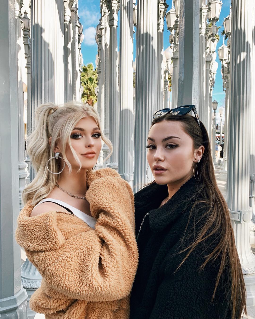 two young women are posing for a portrait in front of the "urban light" art installation at the los angeles county museum of art (lacma). the woman on the left has blonde hair styled in a high ponytail, a tan teddy bear jacket, and hoop earrings. the woman on the right has long brown hair, sunglasses on her head, a black jacket, and hoop earrings. they are standing among the rows of white columns with street lamps, and a blue sky with some clouds can be seen through the columns.