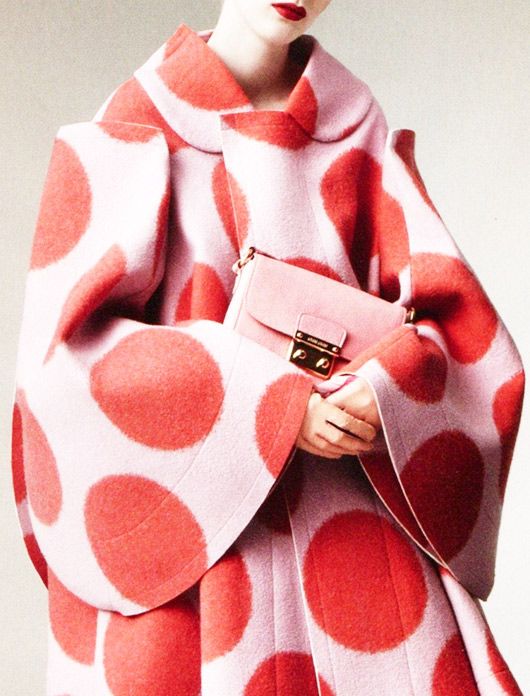 closeup view of a woman modeling a stylish, oversized coat. the coat is predominantly pale pink with large, vibrant red polka dots. it features wide, bellshaped sleeves and a high collar. the model is holding a pale pink handbag, which complements the coat's colors. the overall aesthetic is highfashion, luxurious, and suggestive of autumn or winter attire. the image is sharply focused and has a clean, editorial style reminiscent of fashion magazines like vogue.