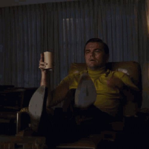 the image shows leonardo dicaprio as rick dalton in the movie "once upon a time in hollywood." he is sitting in a brown leather armchair in a living room, wearing a yellow turtleneck and black pants. he is holding a cup in his hand, looking shocked or surprised. the image is often used as a meme.