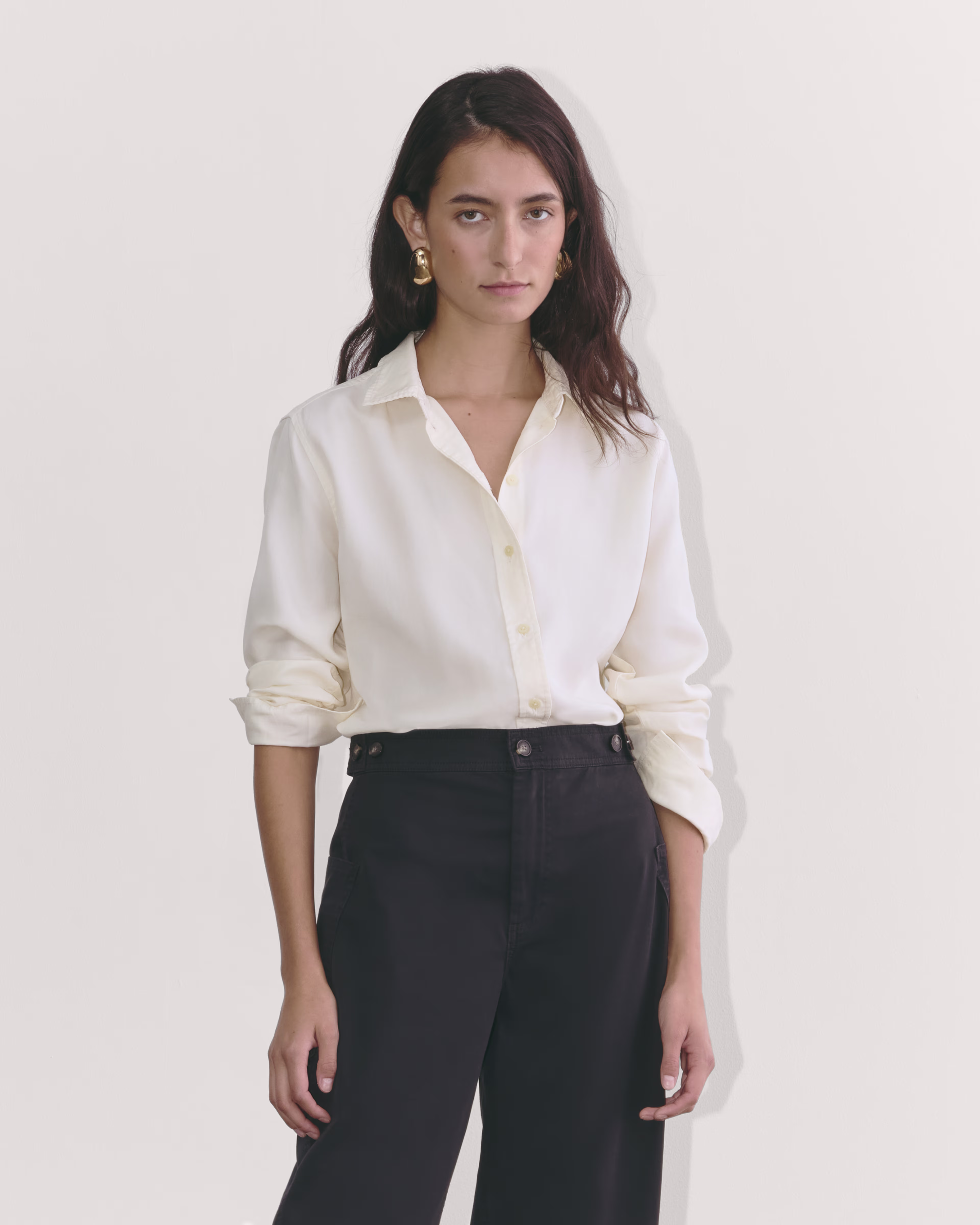 a woman in a white button up shirt and black high waisted pants poses in a studio setting. she has long brown hair and is wearing gold hoop earrings. she is looking directly at the camera.