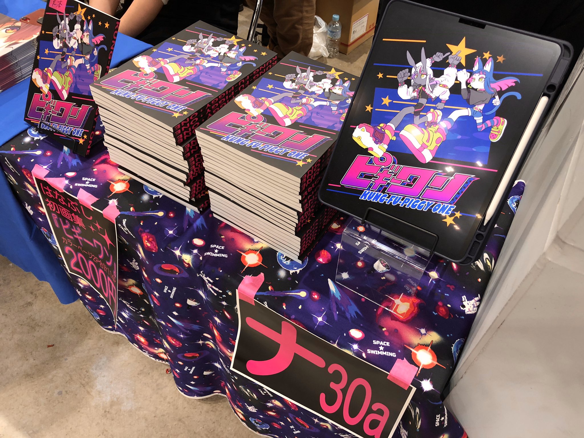 a table with stacks of fanmade manga or comic books featuring colorful characters and a cosmic theme. the table is draped in a black tablecloth with a space pattern, and there are signs indicating the price (30a, possibly 3000 yen) and the artist's circle or space number (possibly 200).  the vibrant artwork and signage suggest this is a booth at a comic convention or similar event.