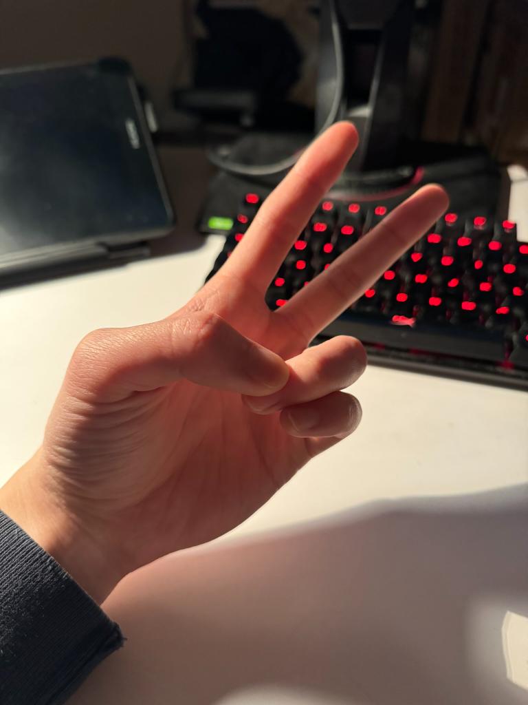 a hand is making a peace sign in front of a dimly lit gaming setup. the hand is in focus while the background is blurred out. the gaming setup includes a laptop, a headset, and a red mechanical keyboard. the photo is taken from a firstperson perspective.