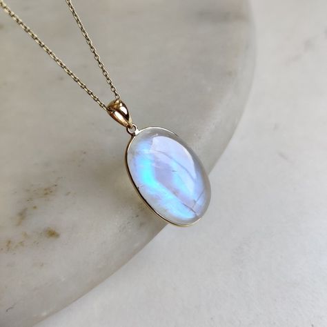 the image shows a gold chain necklace with a large oval moonstone pendant. the moonstone is a pale blue color with a slight iridescence. the necklace is lying on a white marble surface.