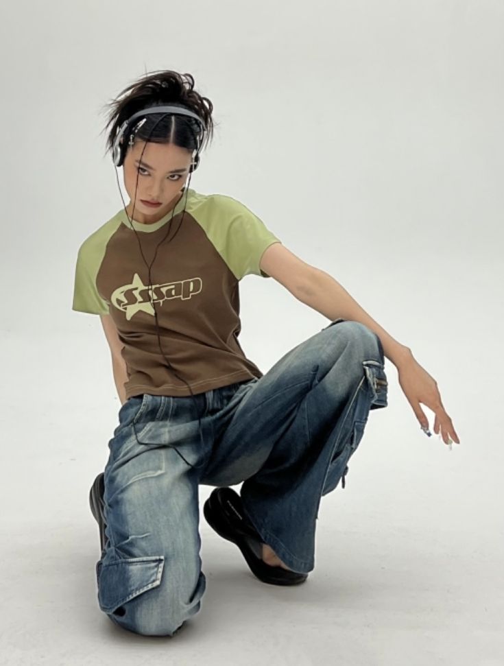 the image shows a young female fashion model posing in a studio against a white background. she's wearing a brown and light green raglansleeved tshirt with a graphic design, and loosefitting, lowrise blue jeans with cargo pockets.  her jeans are baggy and have a wide leg. she's wearing black shoes and has headphones around her neck. the overall style is reminiscent of 2000s/y2k fashion, characterized by its relaxed and casual yet stylish aesthetic.  the model has dark hair and pale skin. her pose is relaxed but striking.