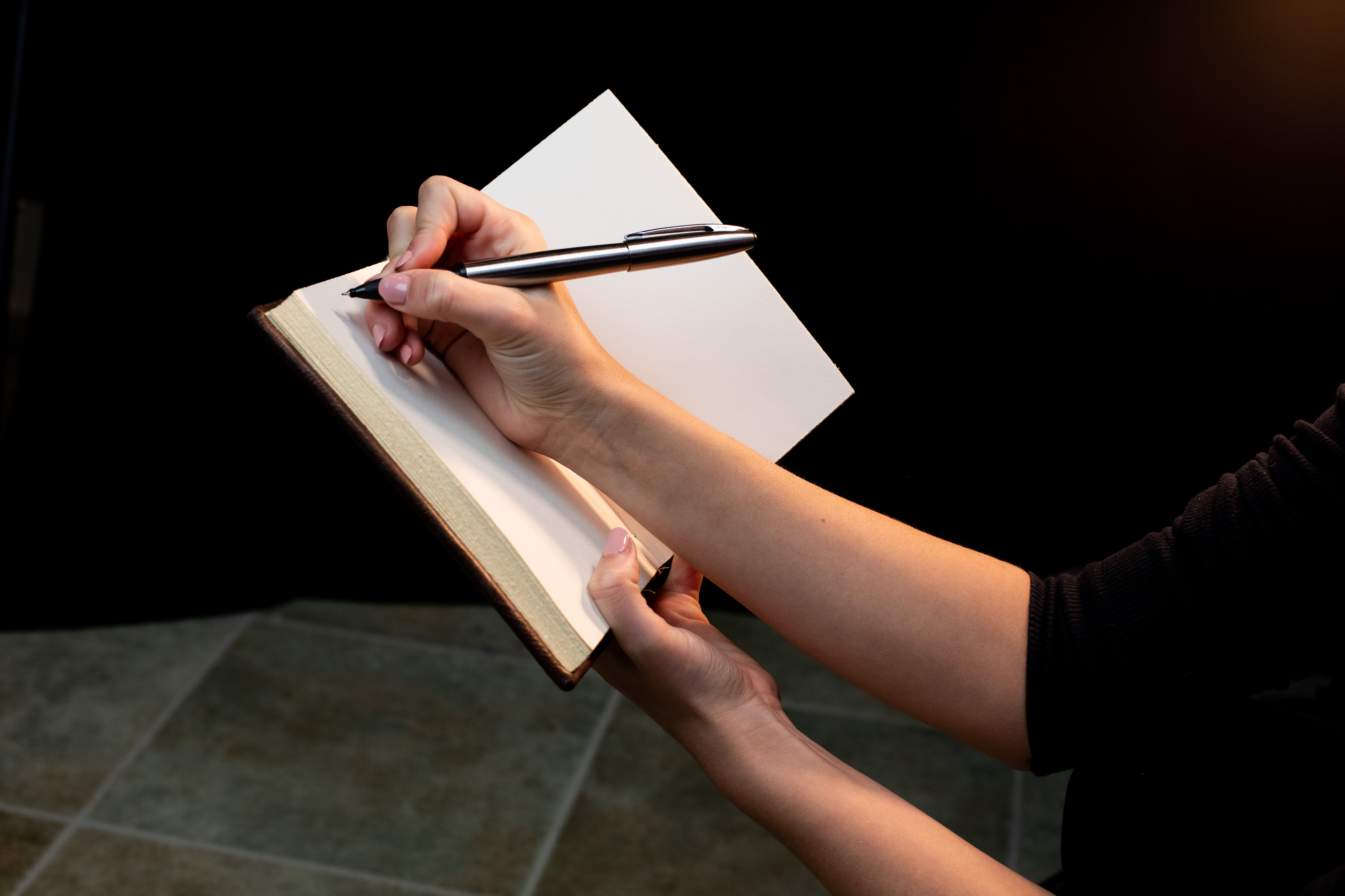 a closeup shot of a woman's hands writing in a leatherbound notebook with a silver pen. the notebook is open to a blank page, and the woman's hand is partially illuminated by a warm light source. the background is dark, creating a sense of intimacy and focus on the act of writing.