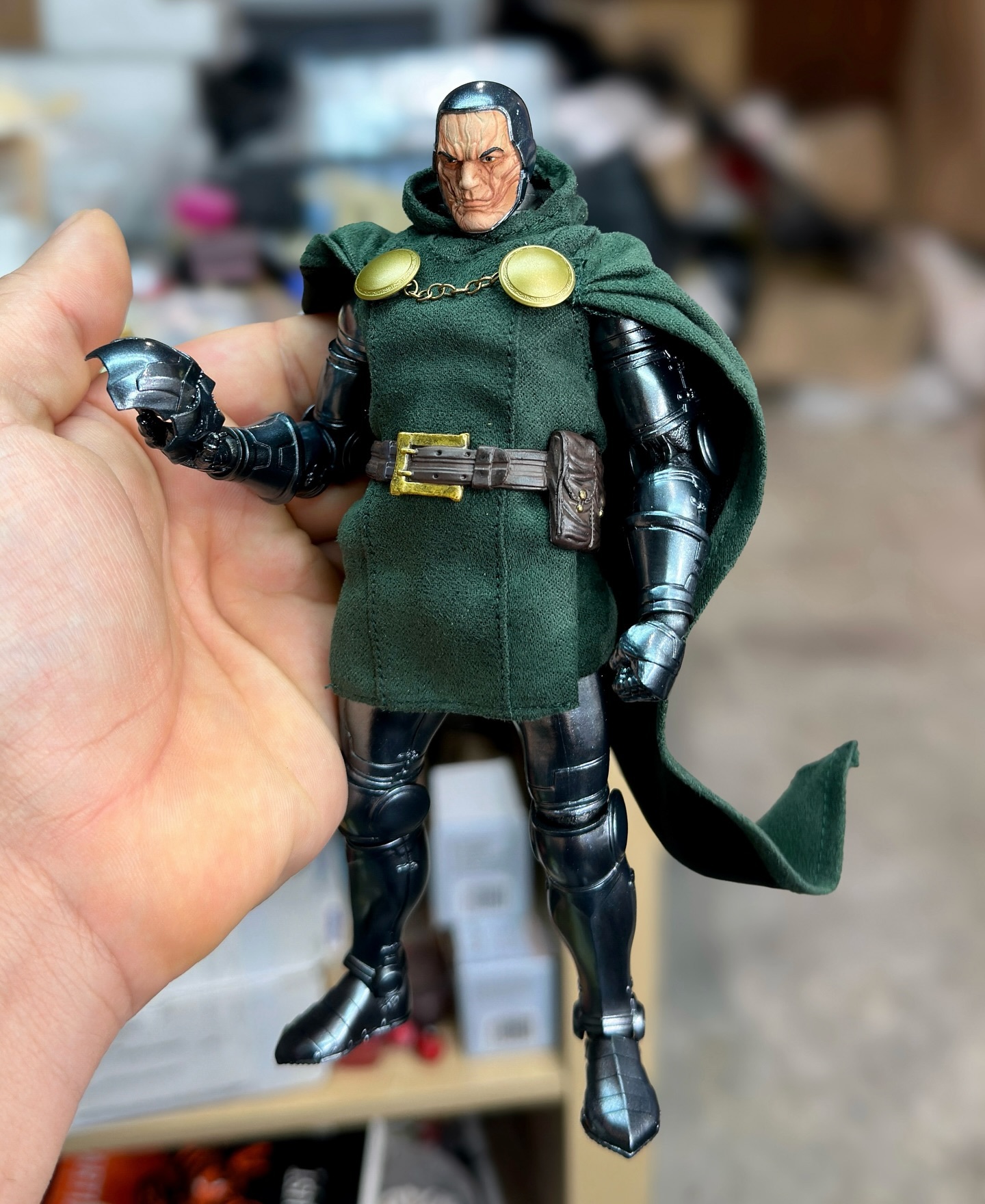 a closeup shot shows a doctor doom action figure being held in someone's hand. the figure is dressed in the character's signature green hooded cloak with golden embellishments and metallic gray armor. he's wearing a belt with a pouch. the figure looks detailed and realistic.