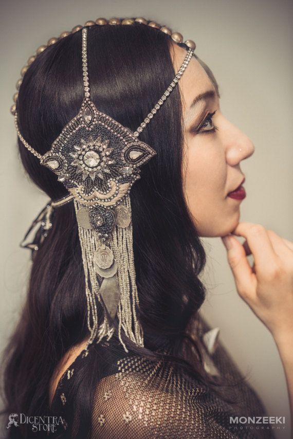 a beautiful young woman with long dark hair, wearing a  silver  and beaded crown headpiece, looks wistfully to her side,  her hand resting on her chin.