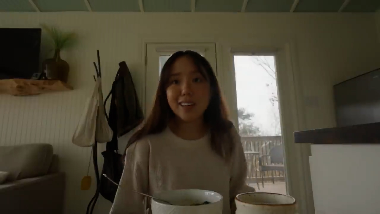 a young woman is sitting at a table in her kitchen, eating a bowl of soup and talking to the camera. she is smiling and appears to be happy and relaxed. the kitchen is cozy and inviting, with warm lighting and neutral colors.
