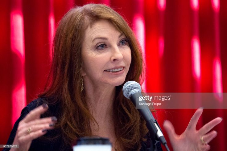 a woman with long brown hair is speaking into a microphone. she is standing on a stage with a red background. she is wearing a black dress and a necklace. she is smiling and appears to be confident.