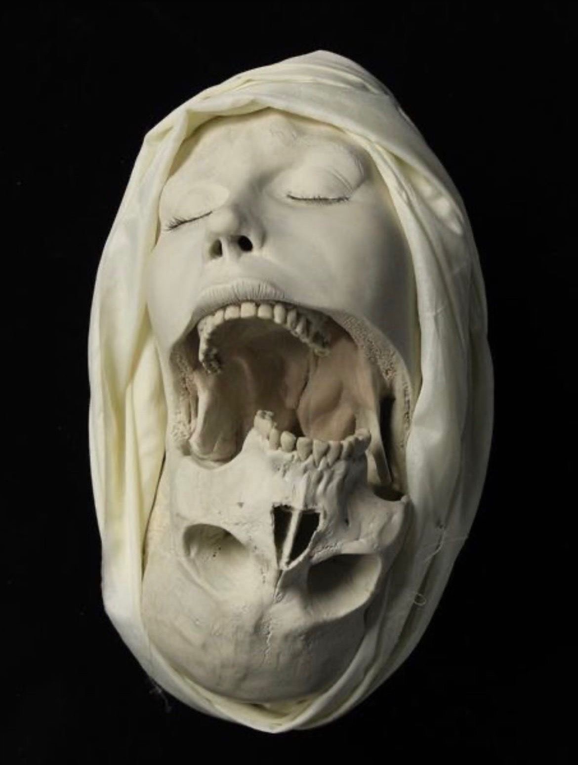 a white sculpture of a woman's head with her mouth open, revealing a skull inside. the woman's eyes are closed, and she is wearing a white head covering. the sculpture is mounted on a black background.