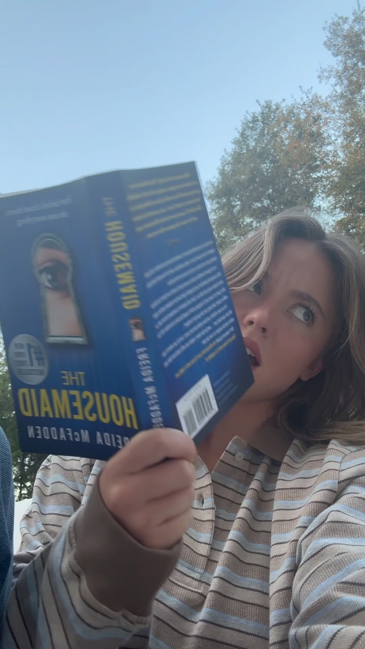 a closeup shot of a young woman, possibly a teenager, sitting outdoors under a partly sunny sky with trees visible in the background. she is wearing a light brown and blue striped longsleeved shirt and is holding and reading a book titled "the housemaid" by frida mcfadden. the book cover is predominantly blue with a keyhole image. the woman's expression is one of concentration or slight concern as she reads. the overall mood is relaxed yet somewhat mysterious due to the book cover.