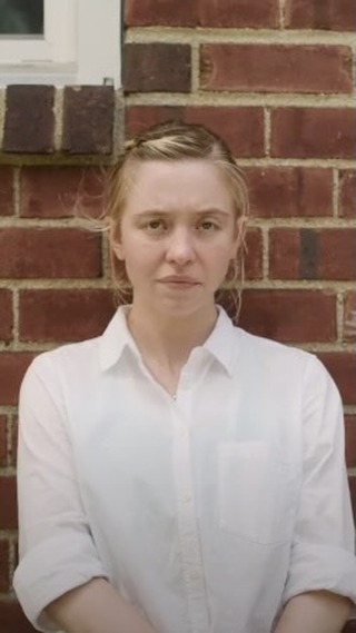the image shows a closeup portrait of a young, lightskinned blonde woman.  she's wearing a simple, white, buttondown shirt and has her hair pulled back from her face.  she is standing against a red brick wall, her expression is neutral and somewhat serious or contemplative. the setting appears to be outdoors in natural light. the overall mood of the image is calm and understated.